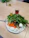 A plate of assorted vegetables and a boiled egg is placed on a wooden table. The plate contains leafy greens, sliced carrots, broccoli, radishes, and assorted olives. Alongside the plate, there are small glasses containing olive oil and a red beverage.