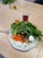 A plate of assorted vegetables and a boiled egg is placed on a wooden table. The plate contains leafy greens, sliced carrots, broccoli, radishes, and assorted olives. Alongside the plate, there are small glasses containing olive oil and a red beverage.