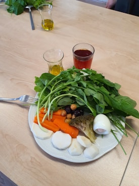 A plate of assorted vegetables and a boiled egg is placed on a wooden table. The plate contains leafy greens, sliced carrots, broccoli, radishes, and assorted olives. Alongside the plate, there are small glasses containing olive oil and a red beverage.