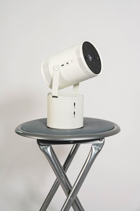 A modern projector with a cylindrical shape rests on a round, metallic folding table. The projector is white with a black lens and several ports on its side. The table has crossed metal legs providing support, placed against a plain light grey background.