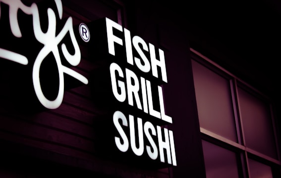 A brightly lit sign with white lettering displaying the words 'FISH GRILL SUSHI' against a dark background. The sign is mounted on a building exterior with some visible windows.