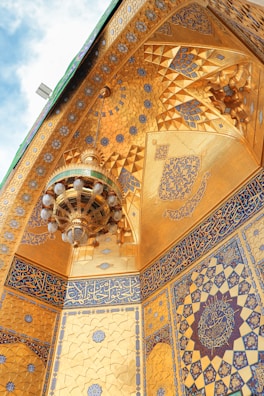 An intricately decorated interior featuring golden walls with geometric patterns, ornate blue arabesque designs, and Arabic calligraphy. A large, elaborate chandelier is suspended from the ceiling, adorned with multiple glass spheres.