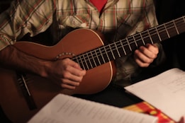 A person is playing an acoustic guitar with a visible plaid shirt. The focus is on the hands and guitar, while sheets of paper are blurred in the foreground.