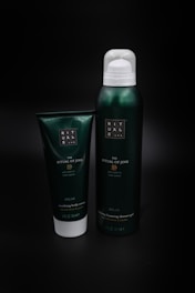 Two green containers are placed against a dark background. The larger one is a metal or plastic canister with a white cap, labeled as 'The Ritual of Jing' foaming shower gel. The smaller container is a tube labeled as soothing body cream.