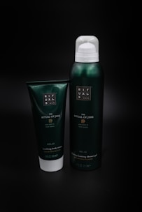 Two green containers are placed against a dark background. The larger one is a metal or plastic canister with a white cap, labeled as 'The Ritual of Jing' foaming shower gel. The smaller container is a tube labeled as soothing body cream.