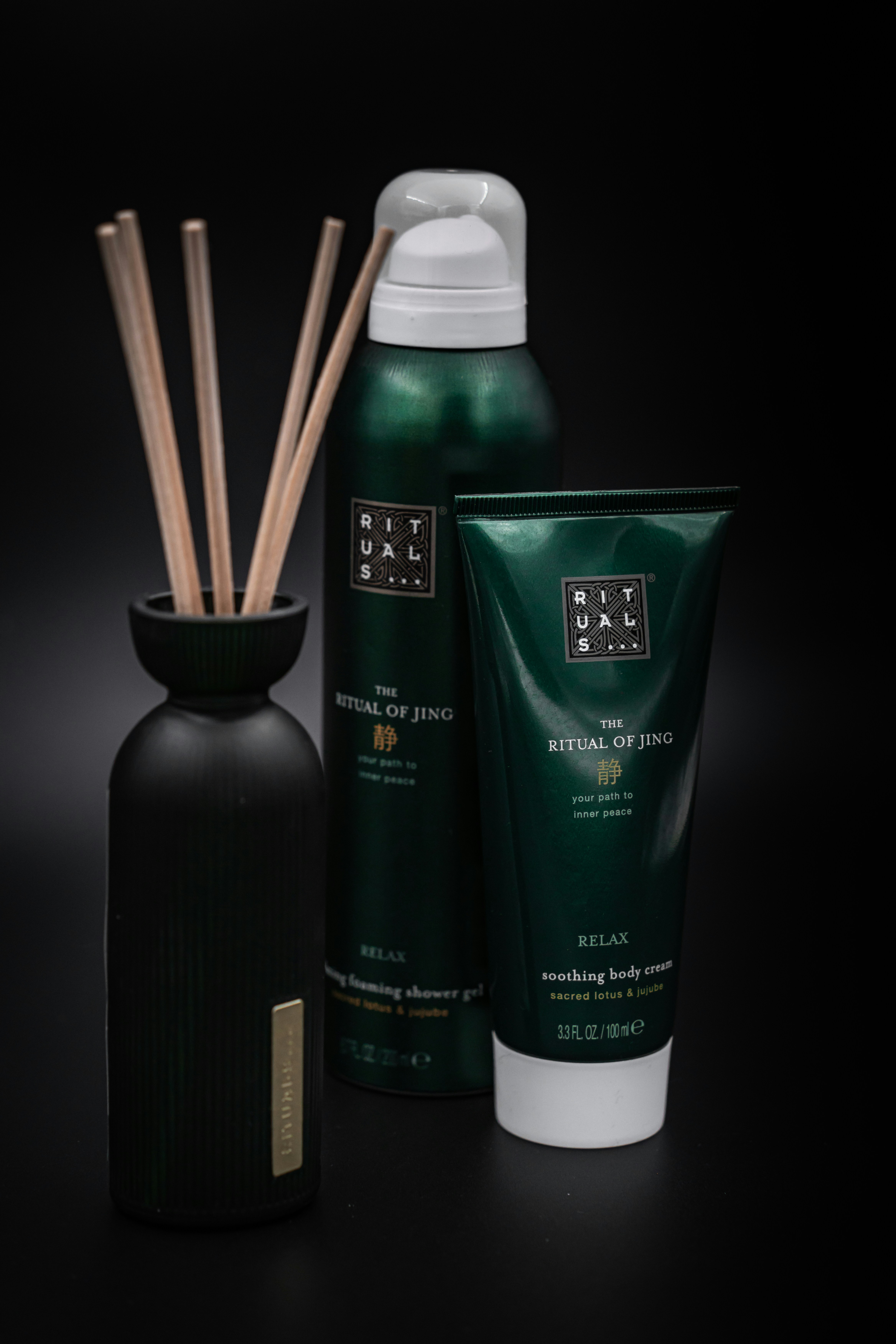 A set of personal care products, including a bottle with reeds, a foaming shower gel, and a soothing body cream tube from the Ritual of Jing collection. The containers are predominantly dark green with minimalist labels, set against a black background that highlights their luxurious appearance.