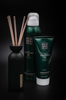 A set of personal care products, including a bottle with reeds, a foaming shower gel, and a soothing body cream tube from the Ritual of Jing collection. The containers are predominantly dark green with minimalist labels, set against a black background that highlights their luxurious appearance.
