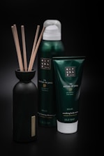A set of personal care products, including a bottle with reeds, a foaming shower gel, and a soothing body cream tube from the Ritual of Jing collection. The containers are predominantly dark green with minimalist labels, set against a black background that highlights their luxurious appearance.