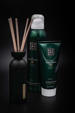 A set of personal care products, including a bottle with reeds, a foaming shower gel, and a soothing body cream tube from the Ritual of Jing collection. The containers are predominantly dark green with minimalist labels, set against a black background that highlights their luxurious appearance.