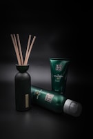 A collection of green and black personal care products is arranged against a dark background. A bottle with wooden sticks is positioned on the left, while two tubes of different sizes are lying and standing to its right.