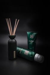 A collection of green and black personal care products is arranged against a dark background. A bottle with wooden sticks is positioned on the left, while two tubes of different sizes are lying and standing to its right.