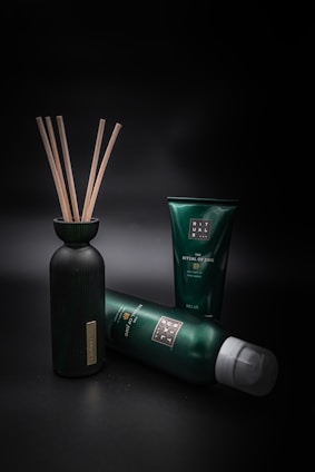 A collection of green and black personal care products is arranged against a dark background. A bottle with wooden sticks is positioned on the left, while two tubes of different sizes are lying and standing to its right.