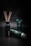 A collection of green and black personal care products is arranged against a dark background. A bottle with wooden sticks is positioned on the left, while two tubes of different sizes are lying and standing to its right.