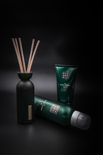 A collection of green and black personal care products is arranged against a dark background. A bottle with wooden sticks is positioned on the left, while two tubes of different sizes are lying and standing to its right.