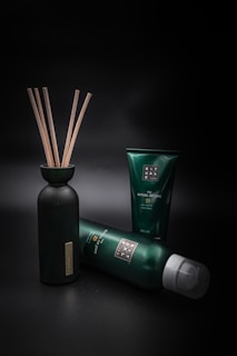 A collection of green and black personal care products is arranged against a dark background. A bottle with wooden sticks is positioned on the left, while two tubes of different sizes are lying and standing to its right.