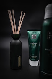 Three products are displayed against a dark, blurred background. On the left is a black bottle with wooden sticks protruding from its opening. In the center is a green tube labeled 'The Ritual of Jing' and described as 'Nourishing Body Cream' with additional text. On the right is part of another bottle with similar branding but the exact details are not visible.