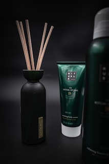 Three products are displayed against a dark, blurred background. On the left is a black bottle with wooden sticks protruding from its opening. In the center is a green tube labeled 