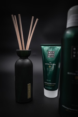Three products are displayed against a dark, blurred background. On the left is a black bottle with wooden sticks protruding from its opening. In the center is a green tube labeled 'The Ritual of Jing' and described as 'Nourishing Body Cream' with additional text. On the right is part of another bottle with similar branding but the exact details are not visible.