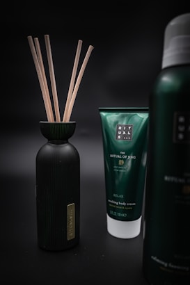 Three products are displayed against a dark, blurred background. On the left is a black bottle with wooden sticks protruding from its opening. In the center is a green tube labeled 'The Ritual of Jing' and described as 'Nourishing Body Cream' with additional text. On the right is part of another bottle with similar branding but the exact details are not visible.