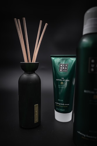Three products are displayed against a dark, blurred background. On the left is a black bottle with wooden sticks protruding from its opening. In the center is a green tube labeled 'The Ritual of Jing' and described as 'Nourishing Body Cream' with additional text. On the right is part of another bottle with similar branding but the exact details are not visible.