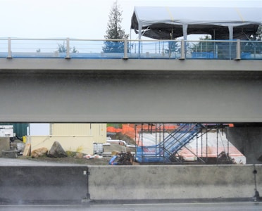 A construction site under a bridge featuring a partially completed structure with scaffolding. There are several construction materials, such as rocks, bags, and tanks, scattered across the ground. An upper section of the bridge includes a canopy with metal railings. Background includes trees and protective orange barriers.