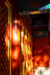 Intricate red and gold lacquered surface with ornate patterns and latticework catches the eye. The sunlight casts dynamic shadows against the structure, highlighting the detailed craftsmanship. Rich colors and detailed carvings emphasize the architectural elegance.