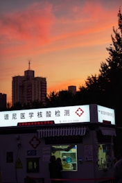 A COVID-19 testing kiosk with a brightly lit sign in Chinese stands in the foreground. The sky is painted with vibrant hues of orange and pink as the sun sets, silhouetting nearby buildings and trees.