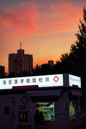 A COVID-19 testing kiosk with a brightly lit sign in Chinese stands in the foreground. The sky is painted with vibrant hues of orange and pink as the sun sets, silhouetting nearby buildings and trees.
