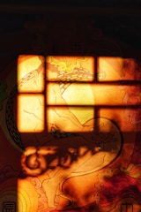 A warm-toned abstract image featuring a silhouette-like form of a person in a seated meditative pose against a vivid background. The light creates rectangular patterns, resembling a window pane, casting shadows and highlighting the intricate swirling designs around the figure.