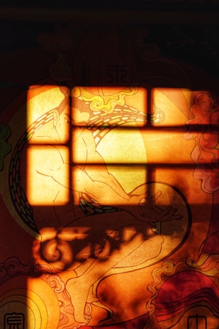 A warm-toned abstract image featuring a silhouette-like form of a person in a seated meditative pose against a vivid background. The light creates rectangular patterns, resembling a window pane, casting shadows and highlighting the intricate swirling designs around the figure.