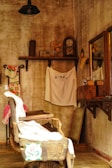 An old-fashioned barbershop with a weathered, rustic aesthetic. The room features a vintage barber chair draped with cloth, a mirror on the wooden wall, and several artifacts like an antique clock and fan on a high shelf. The walls and furniture contribute to a nostalgic, vintage ambiance.