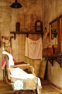 An old-fashioned barbershop with a weathered, rustic aesthetic. The room features a vintage barber chair draped with cloth, a mirror on the wooden wall, and several artifacts like an antique clock and fan on a high shelf. The walls and furniture contribute to a nostalgic, vintage ambiance.