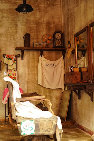 An old-fashioned barbershop with a weathered, rustic aesthetic. The room features a vintage barber chair draped with cloth, a mirror on the wooden wall, and several artifacts like an antique clock and fan on a high shelf. The walls and furniture contribute to a nostalgic, vintage ambiance.