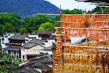 A series of wooden racks holding various dried produce, including corn and other vegetables, are prominently displayed. The background consists of traditional Chinese-style buildings with grey tiled roofs, surrounded by lush green trees. Mountains are visible further in the distance.