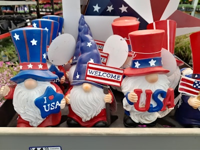 A display of decorative gnome figurines wearing patriotic attire, each featuring a long white beard and oversized hats in red, white, and blue adorned with stars. They hold various signs, such as 'I Love USA', 'Welcome', and other American-themed imagery. The collection appears to be for sale, as indicated by a price tag visible below.
