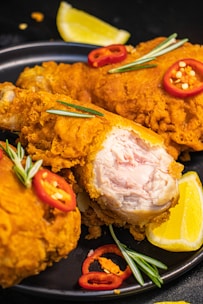 Crispy fried chicken pieces are garnished with red chili slices and rosemary sprigs on a black plate. A wedge of fresh lemon is placed alongside the chicken, adding a pop of color. The texture of the golden-brown batter is evident, with one piece showing the juicy cooked interior.