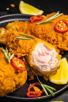 Crispy fried chicken pieces are garnished with red chili slices and rosemary sprigs on a black plate. A wedge of fresh lemon is placed alongside the chicken, adding a pop of color. The texture of the golden-brown batter is evident, with one piece showing the juicy cooked interior.