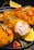 Crispy fried chicken pieces are garnished with red chili slices and rosemary sprigs on a black plate. A wedge of fresh lemon is placed alongside the chicken, adding a pop of color. The texture of the golden-brown batter is evident, with one piece showing the juicy cooked interior.