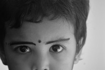 Close-up of a young child's face, focusing on the eyes and eyebrows. The eyebrows are accentuated with dark lines, and there is a small dot on the forehead between the eyebrows. The image is in black and white, highlighting the contrast and details of the facial features.