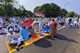 A large group of people gathered outdoors, many sitting on prayer mats in an organized fashion. Several individuals are holding umbrellas, providing shade from the sun. Most people are dressed in traditional attire, wearing caps and robes. The setting is a tree-lined street with a park-like area in the background.