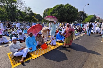 A large group of people gathered outdoors, many sitting on prayer mats in an organized fashion. Several individuals are holding umbrellas, providing shade from the sun. Most people are dressed in traditional attire, wearing caps and robes. The setting is a tree-lined street with a park-like area in the background.