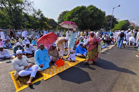 A large group of people gathered outdoors, many sitting on prayer mats in an organized fashion. Several individuals are holding umbrellas, providing shade from the sun. Most people are dressed in traditional attire, wearing caps and robes. The setting is a tree-lined street with a park-like area in the background.
