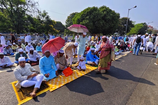 A large group of people gathered outdoors, many sitting on prayer mats in an organized fashion. Several individuals are holding umbrellas, providing shade from the sun. Most people are dressed in traditional attire, wearing caps and robes. The setting is a tree-lined street with a park-like area in the background.
