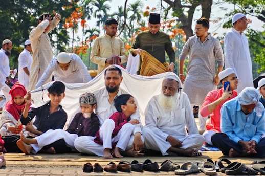 A group of people, including children and adults, are gathered outdoors. Most are wearing traditional attire, such as white clothing and caps. They appear to be engaged in a cultural or religious gathering, sitting together on mats, some under a makeshift cover. The background shows trees and a sunny environment.