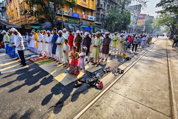 A large group of people is gathered in a street, participating in a prayer session. They are standing on mats, with shoes placed beside them. The scene is set in a city area with buildings and tram tracks visible in the background. The participants wear traditional and colorful attire, including caps.