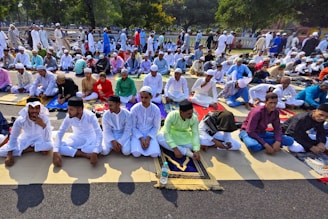 A large group of people are seated outdoors on prayer mats, engaging in a communal prayer ceremony. Most individuals are dressed in traditional attire, including white garments and head coverings. The gathering is taking place on a sunny day with trees visible in the background, suggesting an open public space.