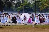A large group of people are seated outdoors on mats, engaged in prayer. Most individuals are dressed in white clothing and wearing traditional caps. The setting is a park or open area surrounded by trees, with some people holding umbrellas for shade.