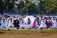 A large group of people are seated outdoors on mats, engaged in prayer. Most individuals are dressed in white clothing and wearing traditional caps. The setting is a park or open area surrounded by trees, with some people holding umbrellas for shade.