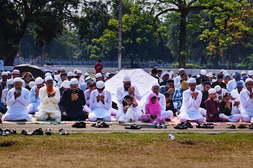 A large group of people are seated outdoors on mats, engaged in prayer. Most individuals are dressed in white clothing and wearing traditional caps. The setting is a park or open area surrounded by trees, with some people holding umbrellas for shade.