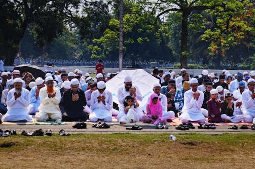 A large group of people are seated outdoors on mats, engaged in prayer. Most individuals are dressed in white clothing and wearing traditional caps. The setting is a park or open area surrounded by trees, with some people holding umbrellas for shade.